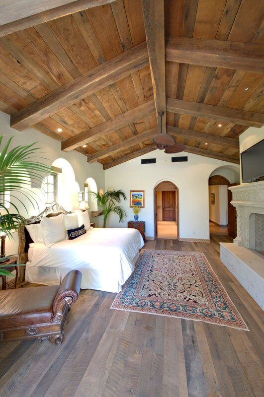 Vaulted bedroom with wooden beams, king-sized bed, cozy leather armchair, tropical plants, and natural light from arched windows.