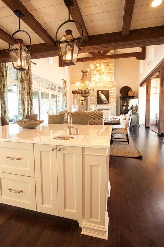 Cozy farmhouse kitchen with white cabinetry, marble island, and natural wood beams. Large windows and decorative fixtures enhance the airy, social atmosphere.