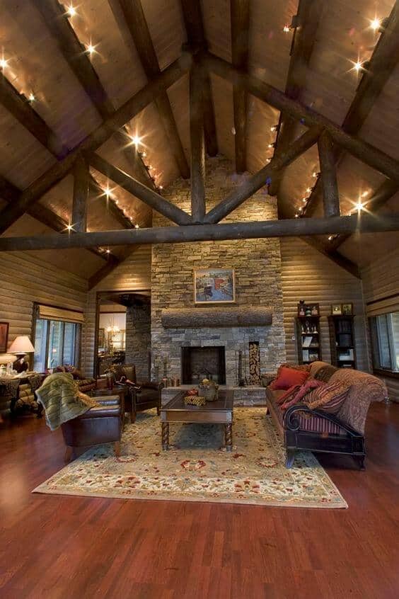 Rustic living room with exposed wooden beams, high ceiling, stone fireplace, floral area rug, leather seating, and natural light.
