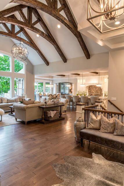 Spacious modern farmhouse living room with exposed wooden beams, light sofas, hardwood flooring, and large windows for natural light.