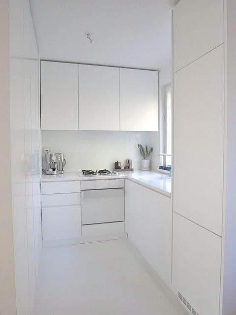 Modern small kitchen with sleek white cabinetry, handleless doors, built-in sink by a window, and spacious white stone countertop.