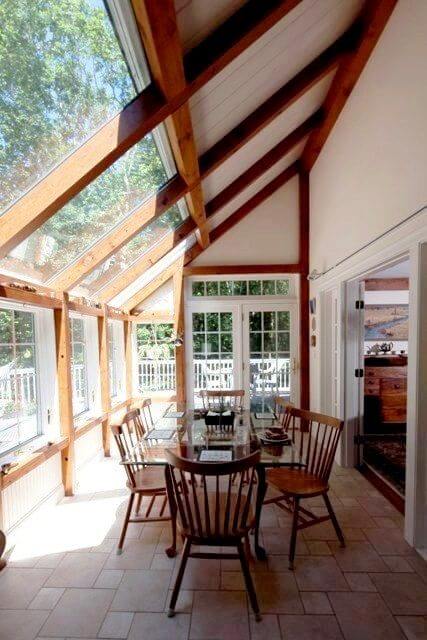 Bright sunroom with large windows, wooden beams, a glass dining table, and classic chairs, promoting a cozy, inviting farmhouse interior design.