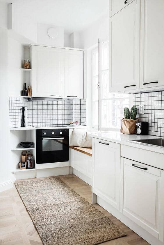 Light-colored cabinetry with black hardware in a corner layout. Polished white quartz countertop, black oven, and geometric backsplash. Bright and airy with large windows, cozy built-in bench, and natural fiber rug. Ideal for small kitchen remodel ideas.