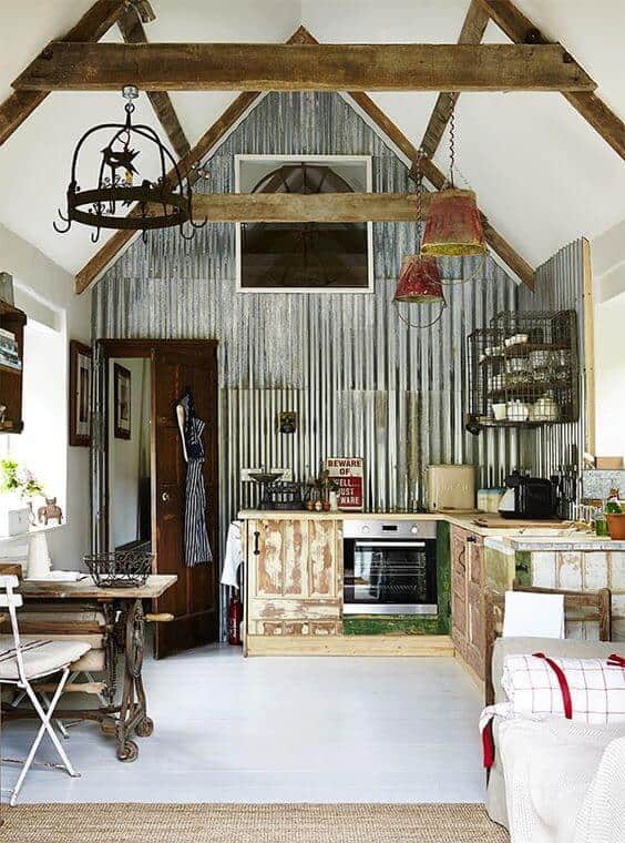 Rustic kitchen with exposed wood beams, corrugated metal wall, wooden island, reclaimed cabinetry, and vintage dining table, brightened by natural light.