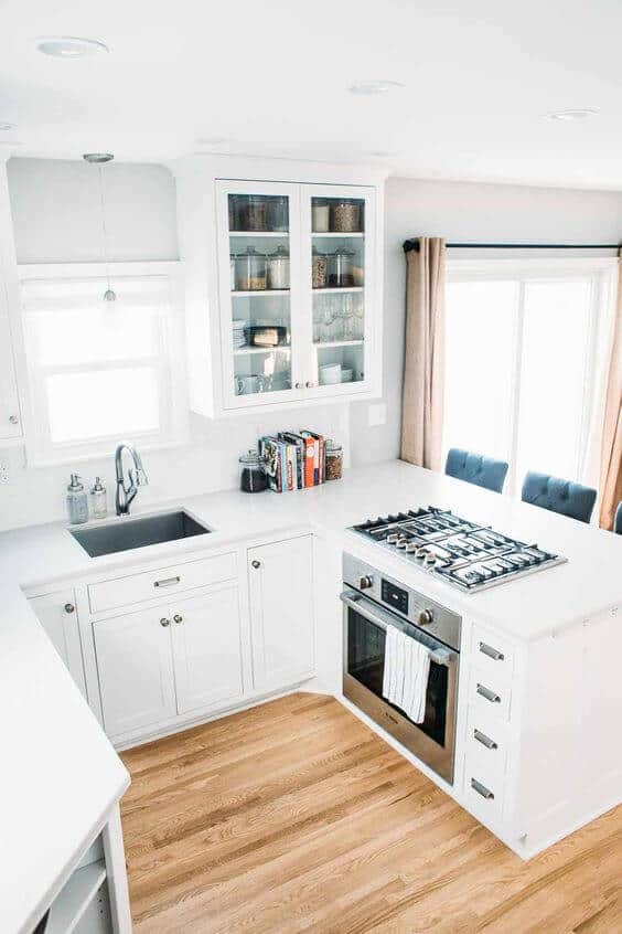 Clean, modern small kitchen with white cabinetry, large countertop, stainless steel sink, and gas cooktop. Glass-front cabinets showcase organized dishware.