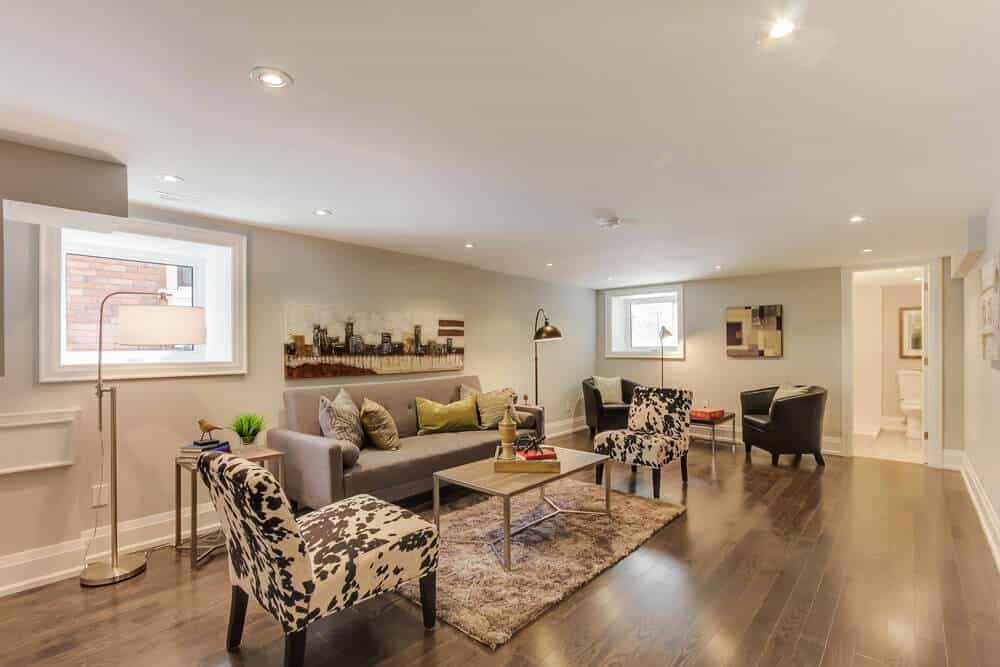 Contemporary basement playroom featuring gray sectional sofa, bold patterned chairs, and plush rug, illuminated by recessed lights and floor lamp.
