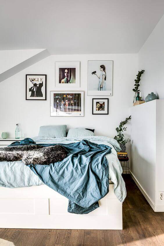 Modern attic bedroom with a queen-sized bed in muted blue hues, eclectic art on white walls, wooden flooring, and natural light.