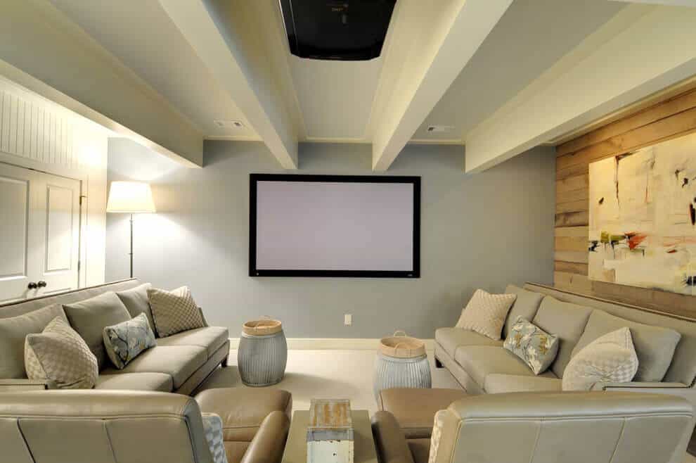Spacious basement playroom with two light gray sofas facing a wall-mounted screen, wood-beam ceiling, and cozy soft gray walls.