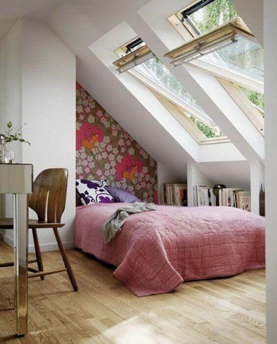 Bright attic bedroom with angled ceiling, queen bed in pink quilt, floral wallpaper, and three skylights. Cozy and functional with bookshelves.