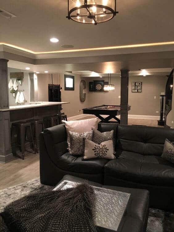 Spacious basement playroom features a dark leather sectional, decorative cushions, and a glass coffee table. Ideal for kids' activities.
