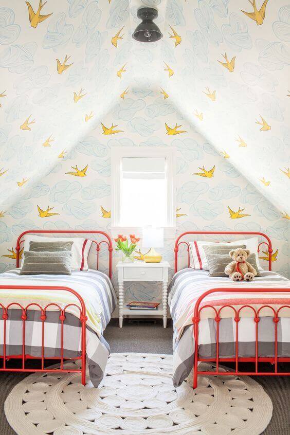 Cozy attic bedroom with two twin beds in red metal frames, sloped ceiling, bird-patterned wallpaper, and a white nightstand.