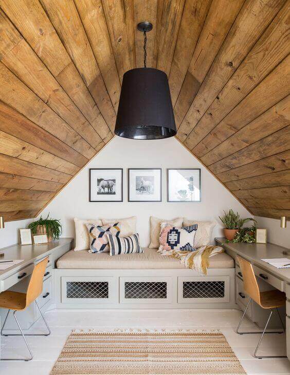 Cozy home office in an attic with a cushioned bench, angled wood paneling, two light-gray desks, and framed artwork.