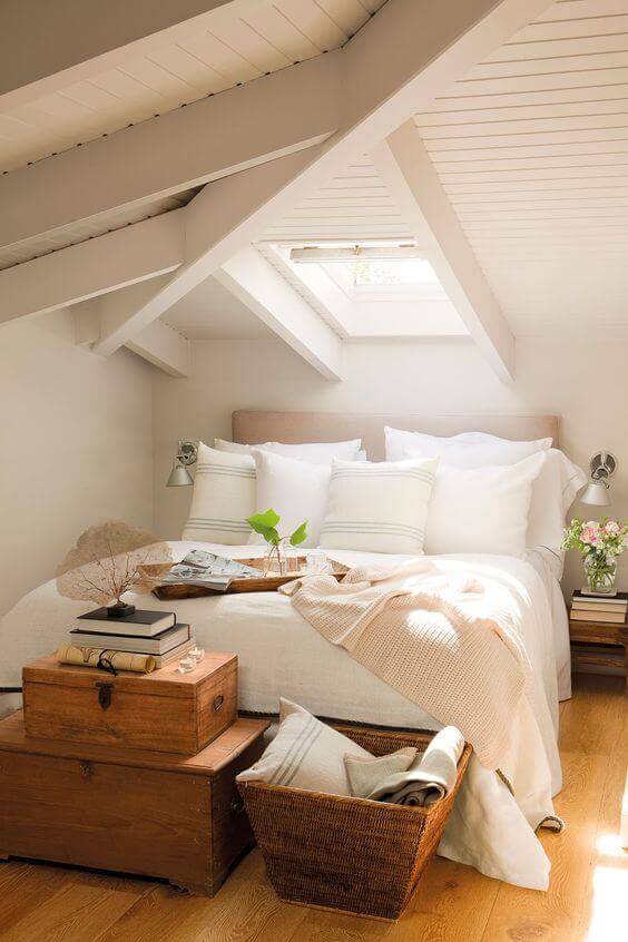 Cozy attic bedroom with sloped ceiling and exposed beams. Features a white duvet, vintage chest, and natural light from a skylight.