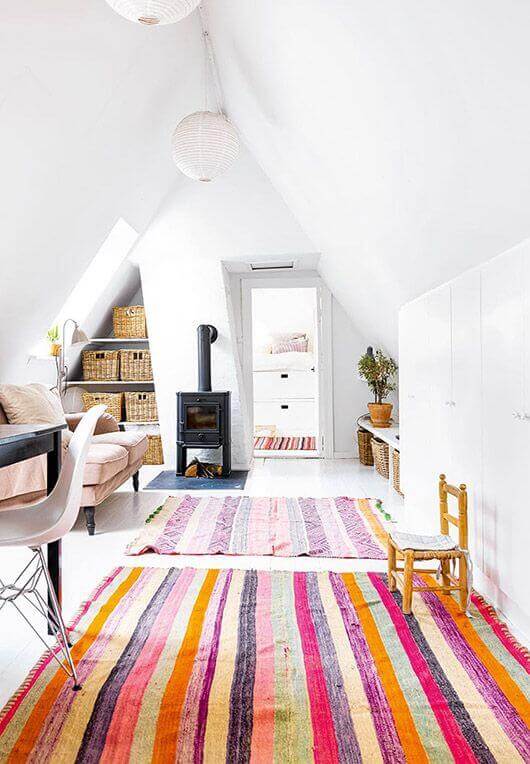 Cozy attic living room with a vibrant striped rug, pink sofa, black desk, and a wood-burning stove. Skylights and pendant lights enhance the airy feel.