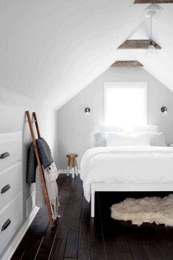Modern bedroom with sloped ceiling and wooden beams. Features a queen-size bed, minimalist stool, and cozy blankets on a ladder. Bright natural light and dark hardwood flooring enhance the design.