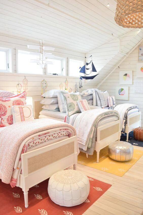 Cozy attic bedroom with two twin beds featuring caned headboards, layered bedding, and colorful pillows. Natural light and nautical decor enhance the inviting atmosphere.