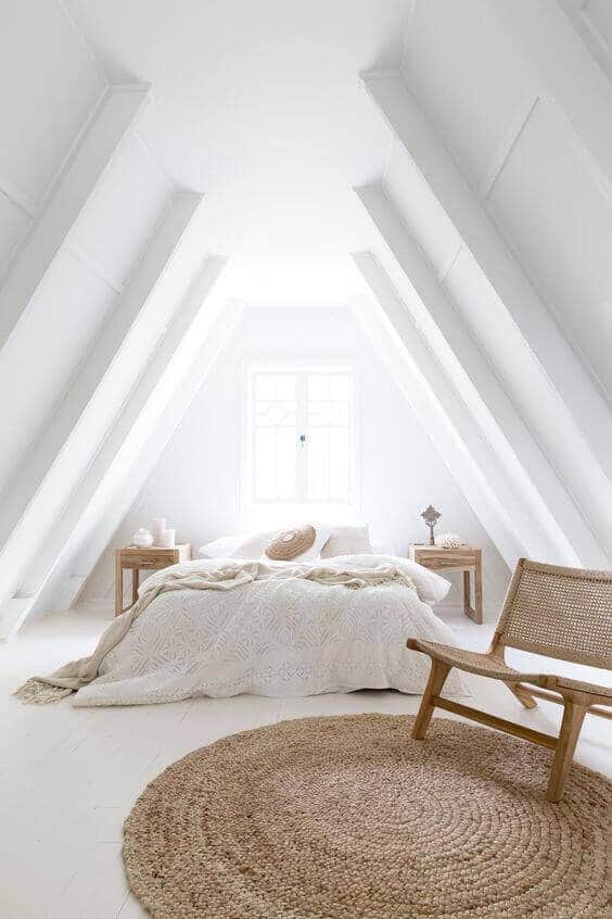 Attic bedroom with sloped white walls, a queen bed with textured bedding, natural light from a large window, and a cozy woven chair.