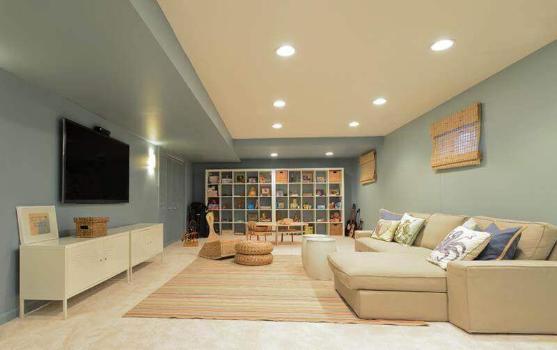Spacious basement playroom with L-shaped sectional sofa, soft blue walls, and modular shelving, creating a cozy, kid-friendly space.