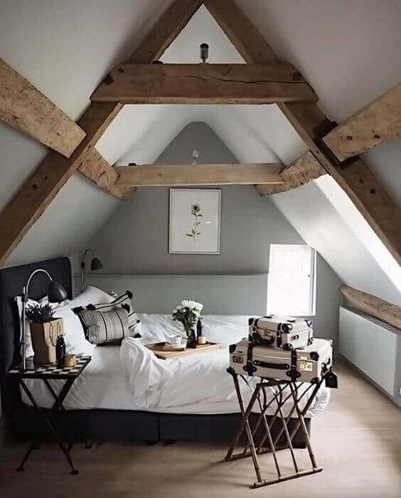 Attic bedroom with sloped ceilings and wooden beams. A centrally placed bed with white linens, accented by striped pillows and a vase of flowers.