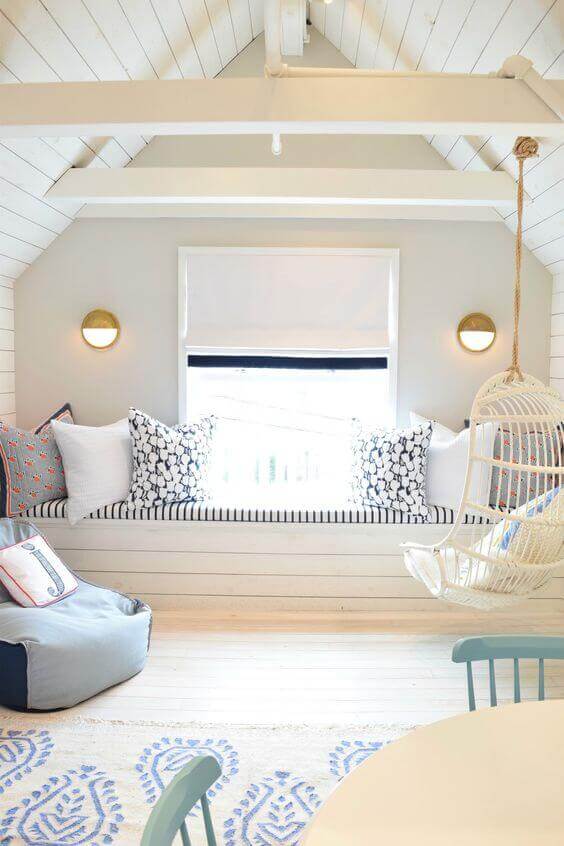 Cozy attic nook with a window seat, cushioned bench, patterned pillows, and a whimsical hanging chair. Light wood flooring enhances the airy vibe.