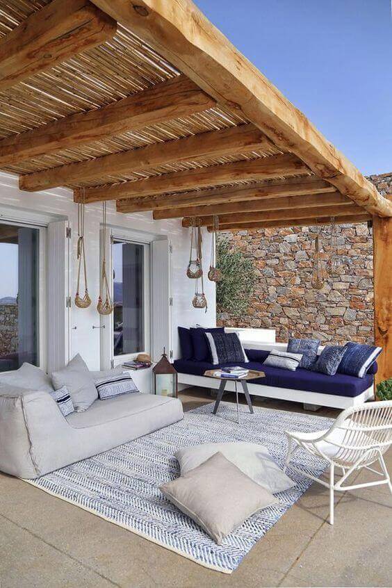 Shaded outdoor living space with wooden beams and thatch roof, featuring light gray and blue sectionals around a coffee table.