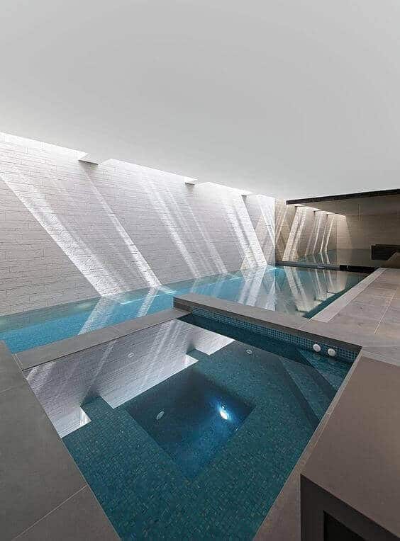 Sleek indoor swim spa featuring a 10x4m water surface, bordered by gray tiles, with a deep blue mosaic spa section and ambient lighting.