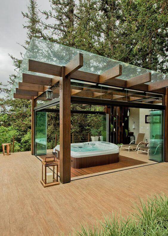 Modern pergola design with a wooden base and glass walls, featuring a sleek hot tub on a spacious deck, surrounded by greenery.
