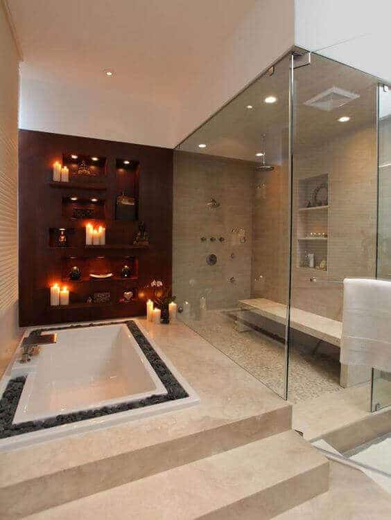Luxury bathroom featuring a freestanding soaking tub surrounded by black pebbles and candles, with a spacious glass-enclosed shower and neutral stone tiles. A wooden shelf displays decor, while a marble bench enhances the minimalist spa vibe.