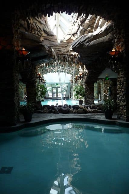 Tranquil indoor swim spa with a curved pool, natural stone walls, and cascading waterfalls, surrounded by lush greenery and warm lighting.