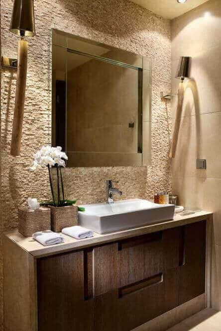 Minimalist spa bathroom featuring textured stone walls, warm lighting, a white sink on a dark wood vanity, and potted greenery.