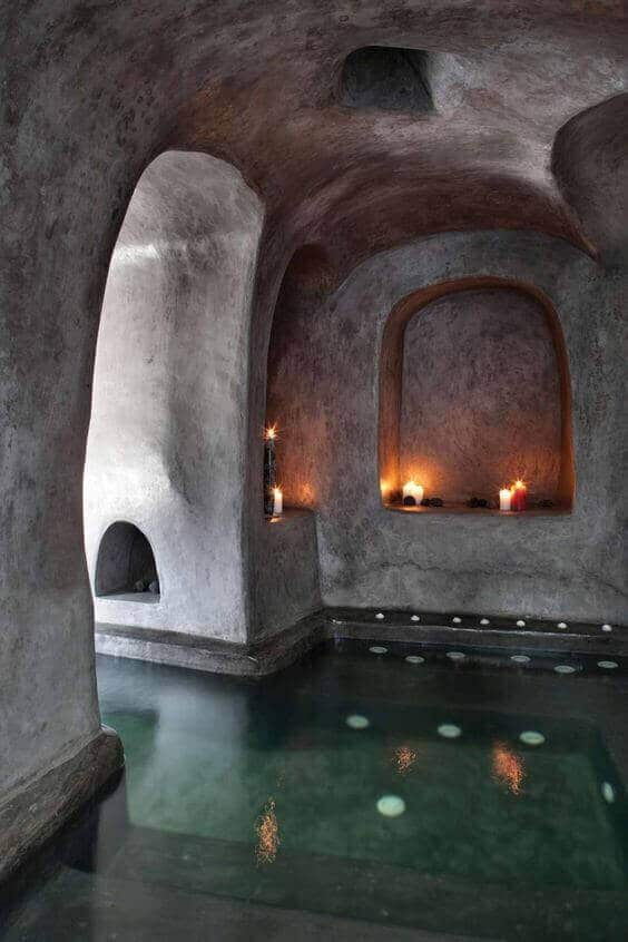 Serene indoor swim spa featuring smooth, curved plaster walls, shallow water area with floating elements, and candle-lit niches.