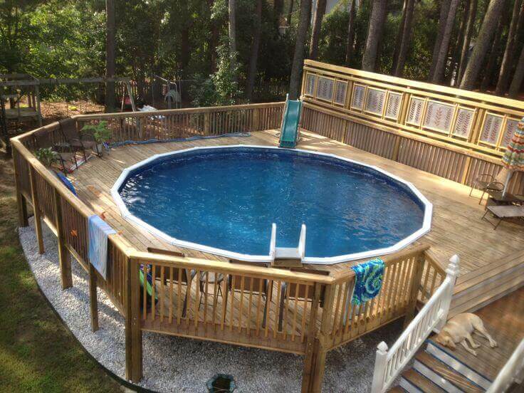 27 Above Ground Pool Landscaping Creative Ideas 13 Above ground pool with a circular design, surrounded by a wooden deck, metal seating, colorful towels, and a blue slide amidst natural greenery.