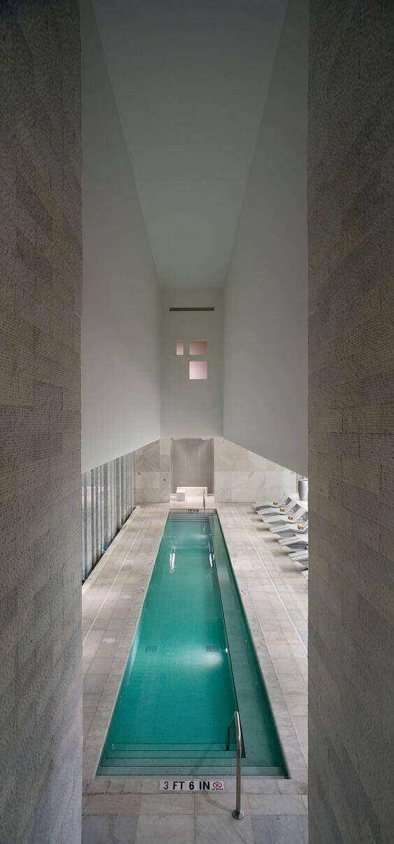 Indoor swim spa features a 10x30 ft design with a glass wall for natural light, light stone walls, and minimalist lounge chairs.