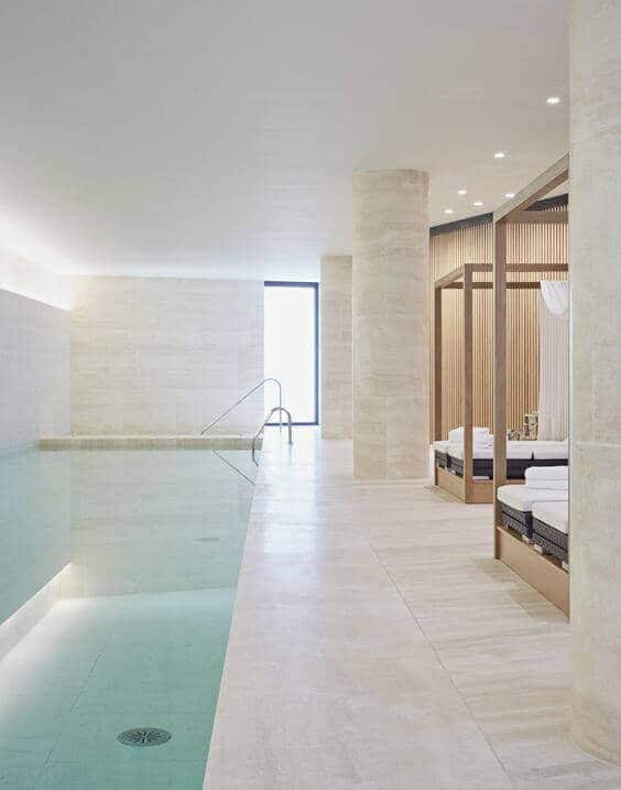 Minimalist indoor swim spa with a 12x4 meter pool, light stone tiles, lounge beds, and large windows for natural light.