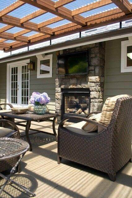 Outdoor patio features a wooden pergola providing partial shade over a woven furniture set and stone fireplace, enhancing relaxation.