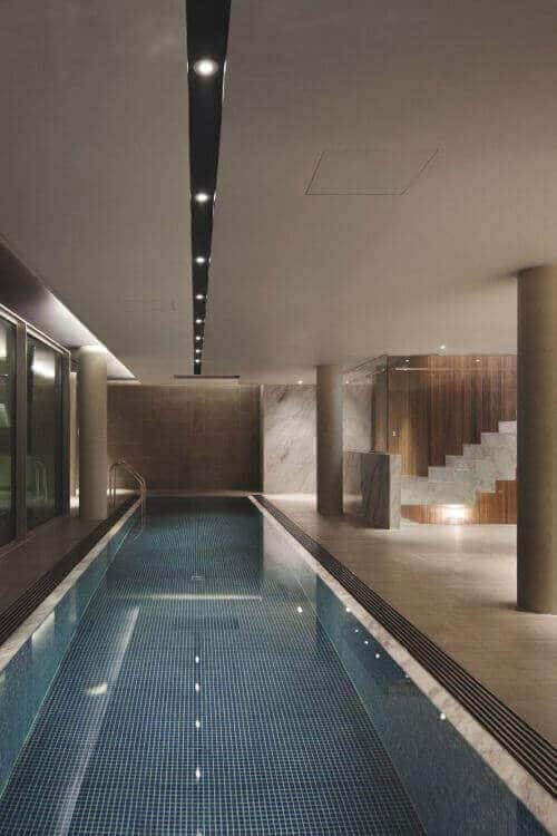 Sleek indoor swim spa featuring large glass panels, dark blue mosaic tiles, and controlled climate for year-round use. Warm wooden accents enhance the space.