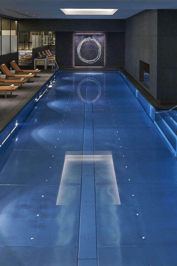 Sleek indoor swim spa featuring a 25-meter azure pool, dark stone walls, ambient lighting, and six wooden lounge chairs for relaxation.