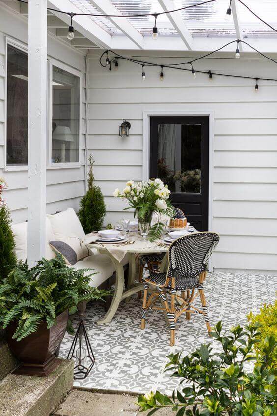 Outdoor dining area with a round wooden table, black and white wicker chairs, and string lights under a transparent roof, accented by greenery.