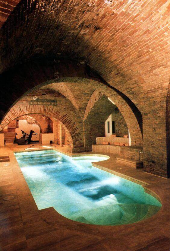 Indoor Swim Spa features a lap pool in a subterranean room, framed by rustic brick arches and stone walls, with cozy seating nearby.