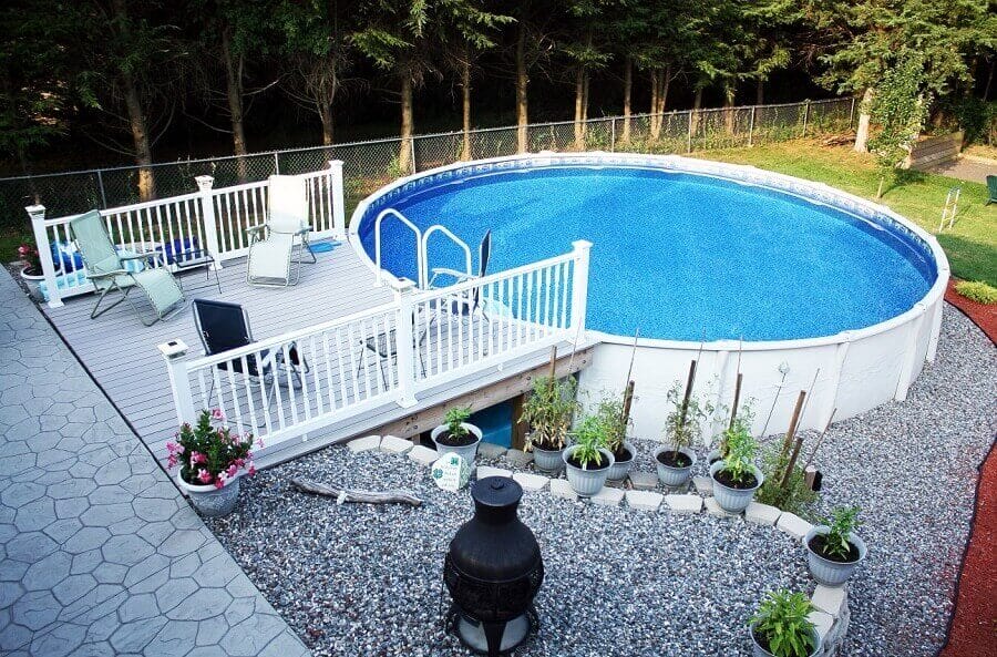 27 Above Ground Pool Landscaping Creative Ideas 18 Round above-ground pool, 24 feet in diameter, on a wooden deck. Surrounded by a white railing, lounge chairs, and potted plants. Green trees provide privacy.