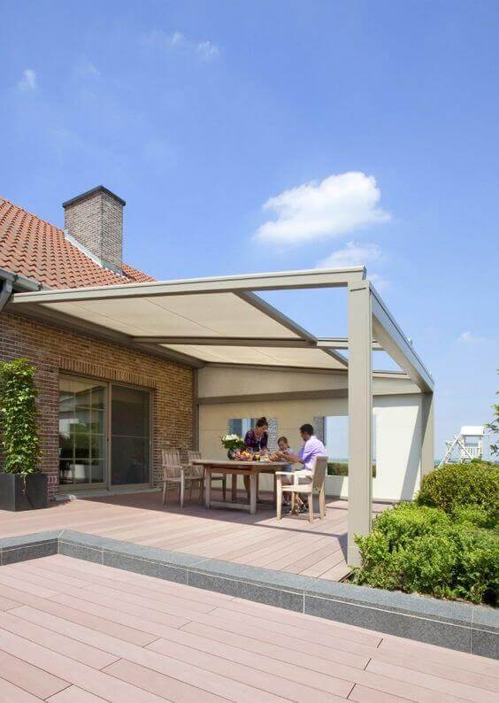 Spacious outdoor dining area with a metal-frame pergola for shade, natural wood table, light chairs, and a lush green hedge for privacy.