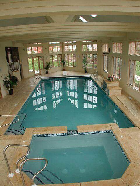 Indoor swim spa features a 20x40 ft rectangular pool with beige tile, adjoining jacuzzi, large windows, wooden beams, and potted plants.