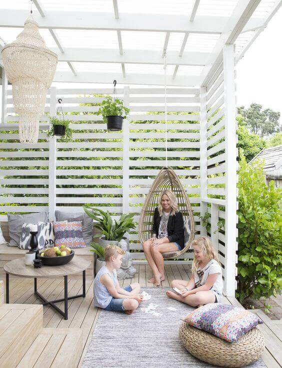 Serene outdoor living space with a light wood deck, white slatted fence, and overhead trellis, featuring an elegant hanging egg chair.