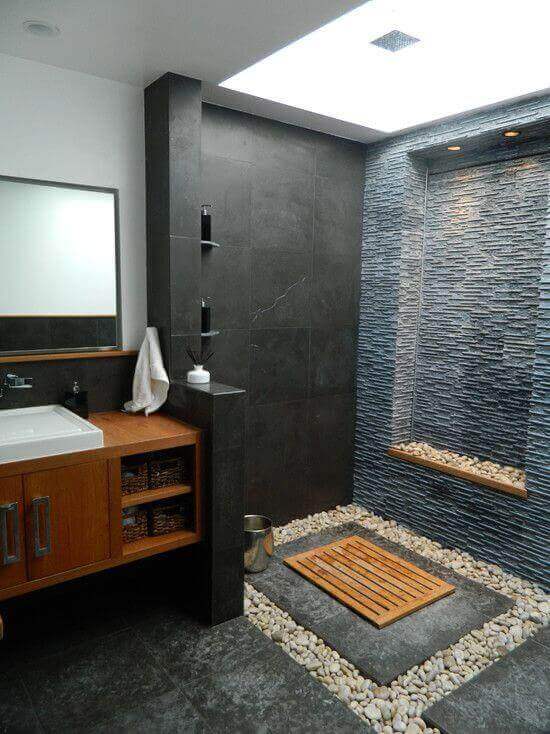Modern spa bathroom with dark stone and wooden accents, open shower, recessed lighting, and pebbled flooring for a serene vibe.