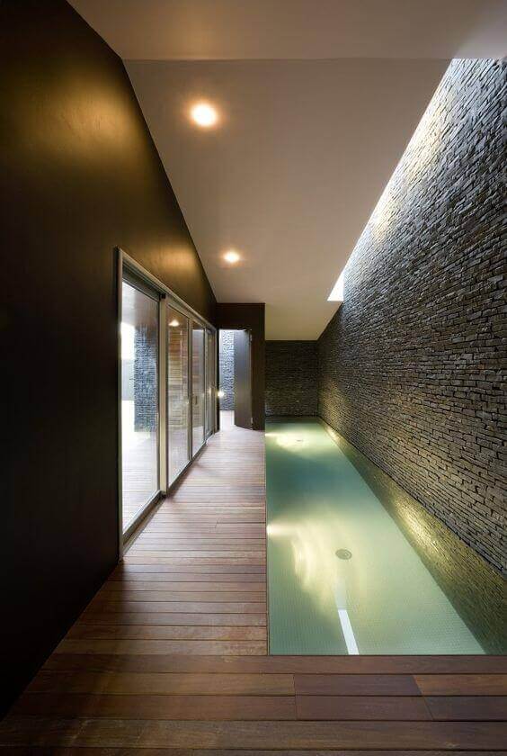 Sleek indoor swim spa features textured stone walls and dark hardwood flooring, illuminated by skylights for a tranquil atmosphere.