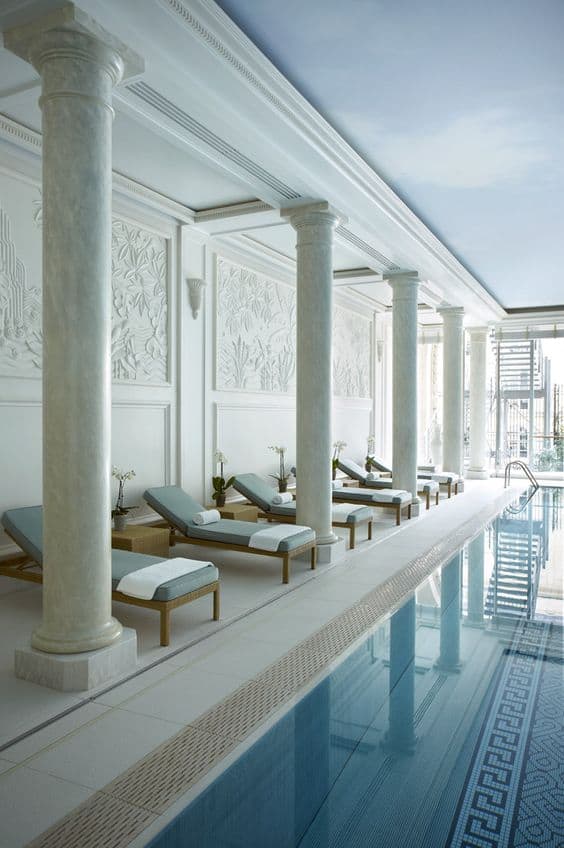 Luxurious indoor swim spa featuring a stunning pool surrounded by tall marble columns, cushioned lounges, and floral-patterned walls.