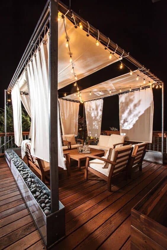Stylish outdoor patio with a metal canopy, flowing fabric curtains, wooden furniture, and ambient string lights, ideal for gatherings.