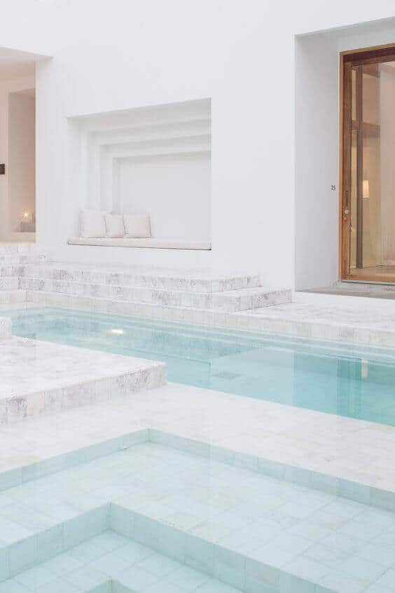 Modern indoor swim spa featuring minimalist design, light marble tiles, and a cozy seating nook, surrounded by natural light.