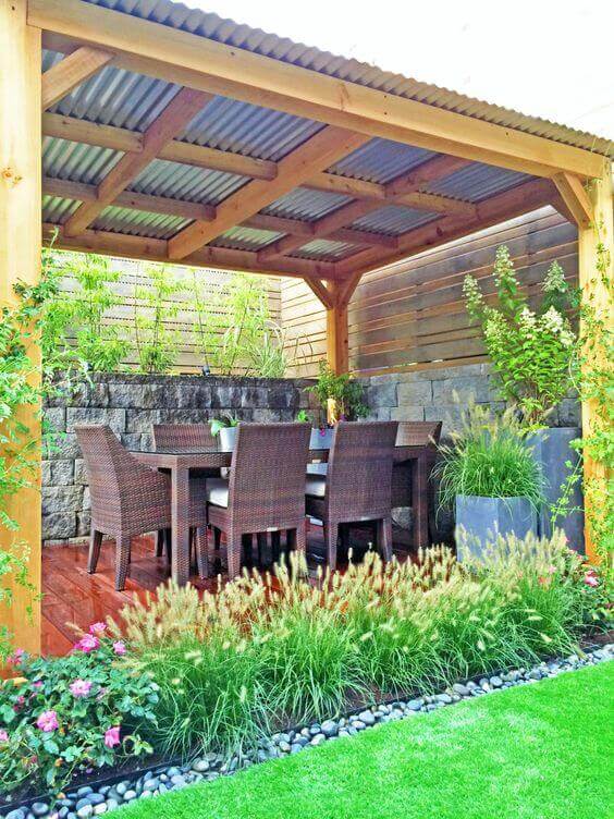 Wooden pergola with a corrugated metal roof shelters a dining area featuring a rectangular table and six wicker chairs, surrounded by greenery.