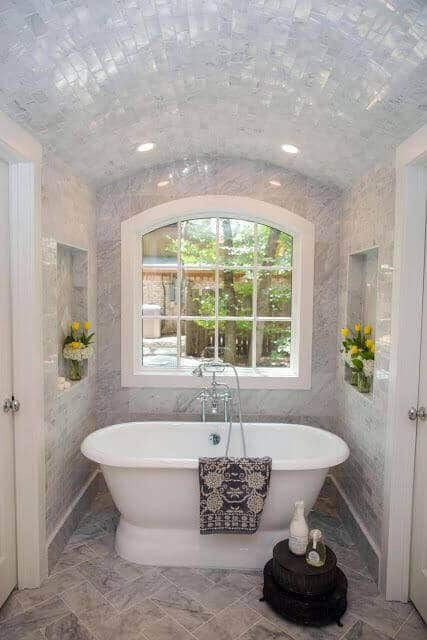 Freestanding oval tub in a spacious bathroom with glossy marble tiles, arched windows, and potted yellow flowers, exuding a spa vibe.